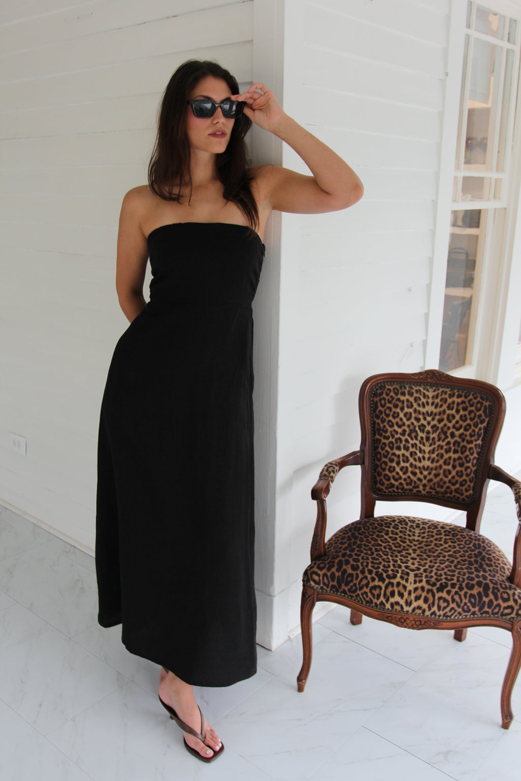 Model wearing a black strapless linen maxi dress with a fitted bodice and straight silhouette, styled with black sandals and sunglasses.