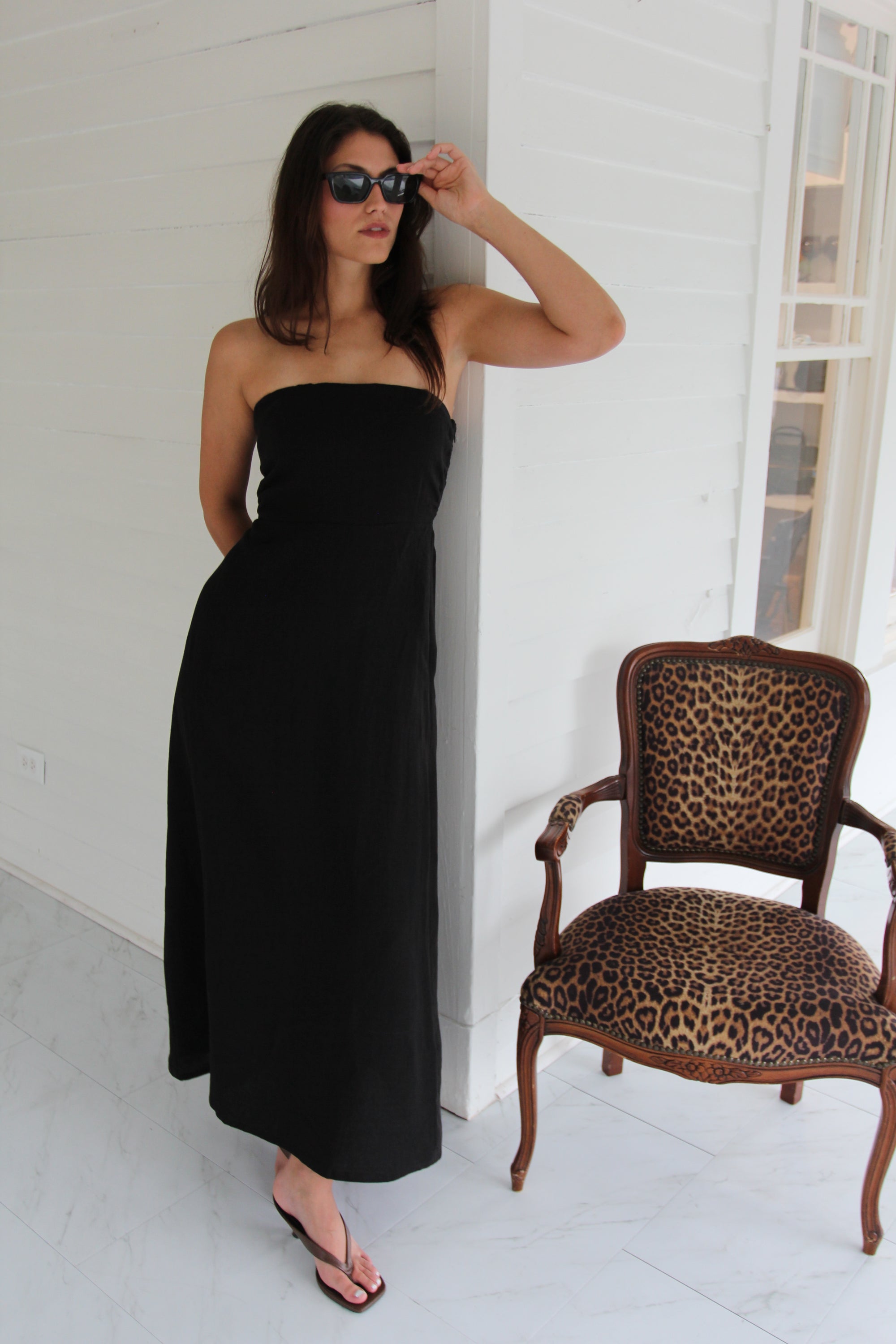 Model wearing a black strapless linen maxi dress with a fitted bodice and straight silhouette, styled with black sandals and sunglasses.