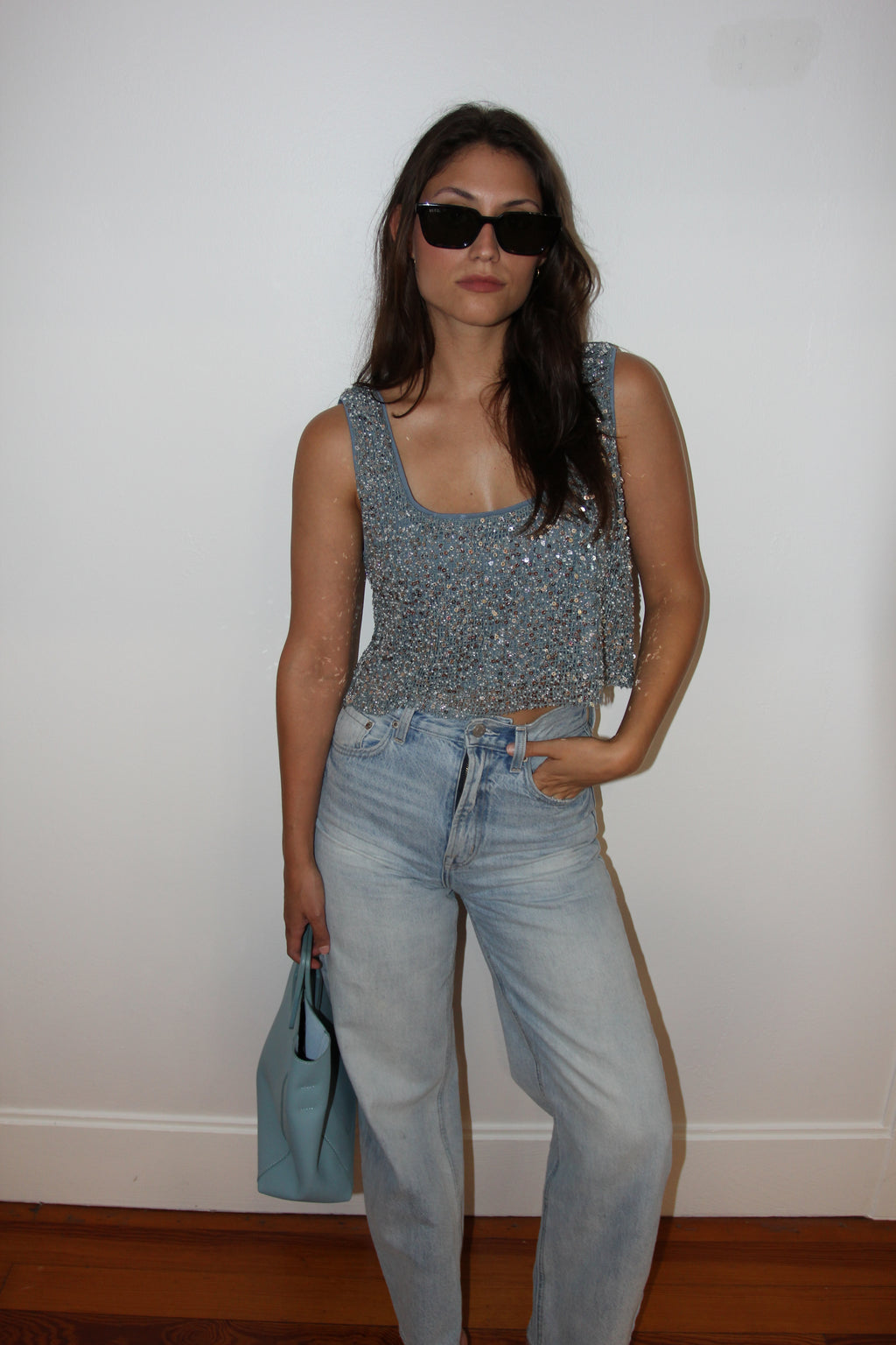 Woman wearing a floral top and jeans, holding a blue handbag against a white wall.