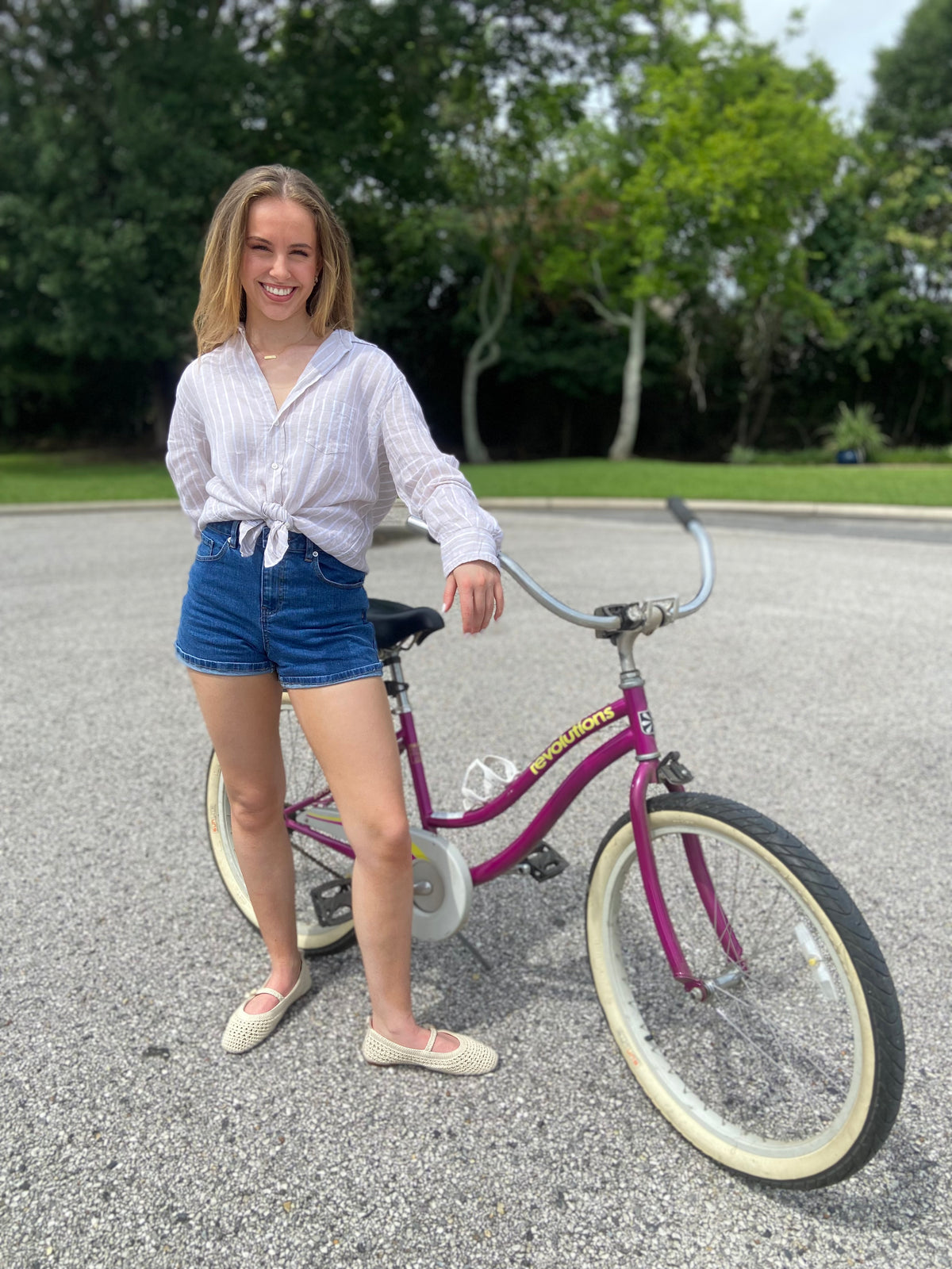 Woman standing next to a purple bicycle on a paved road with trees in the background wearing Frank & Eileen