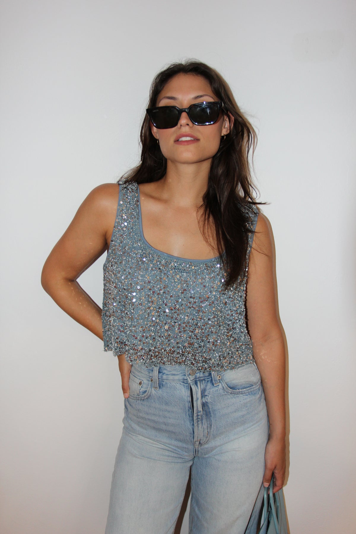 Model wearing a dusty blue sequin crop top with a square neckline, styled with light wash denim and black sunglasses.
