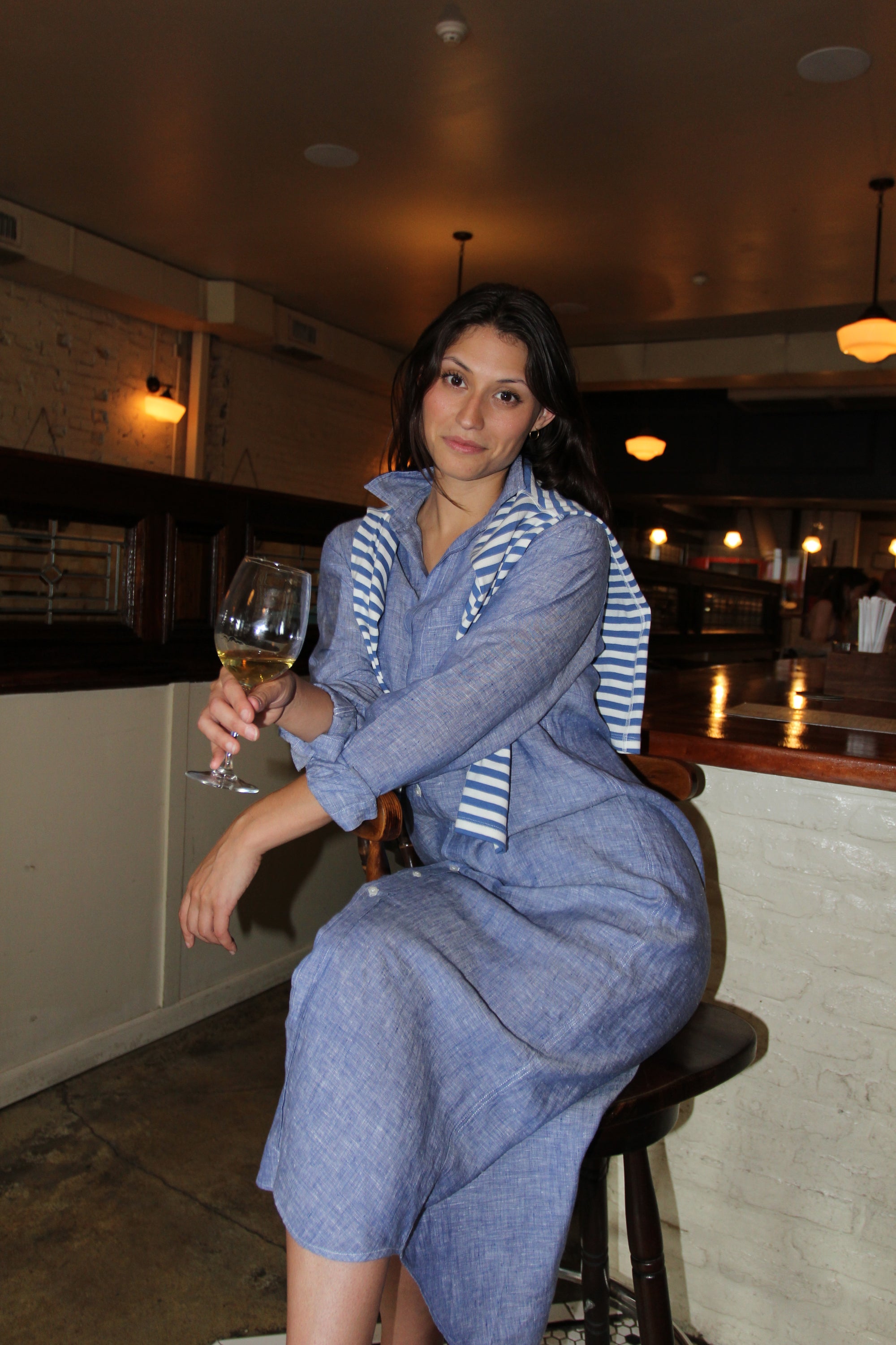 Woman in a blue Italian linen dress sitting on a bar stool holding a glass of wine. Frank & Eileen Rory Dress
