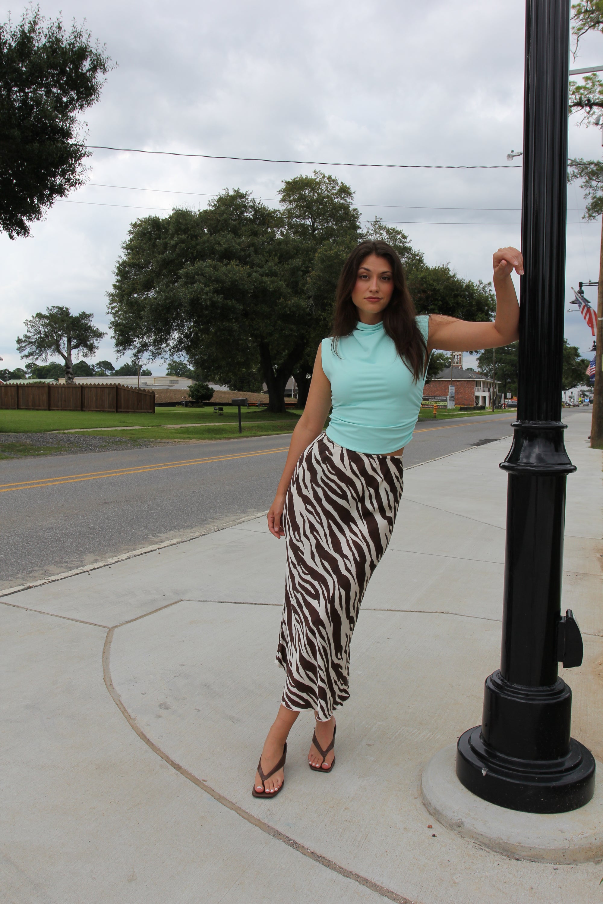Model wearing a brown and ivory zebra print satin midi skirt styled with a turquoise sleeveless high-neck top and brown sandals.